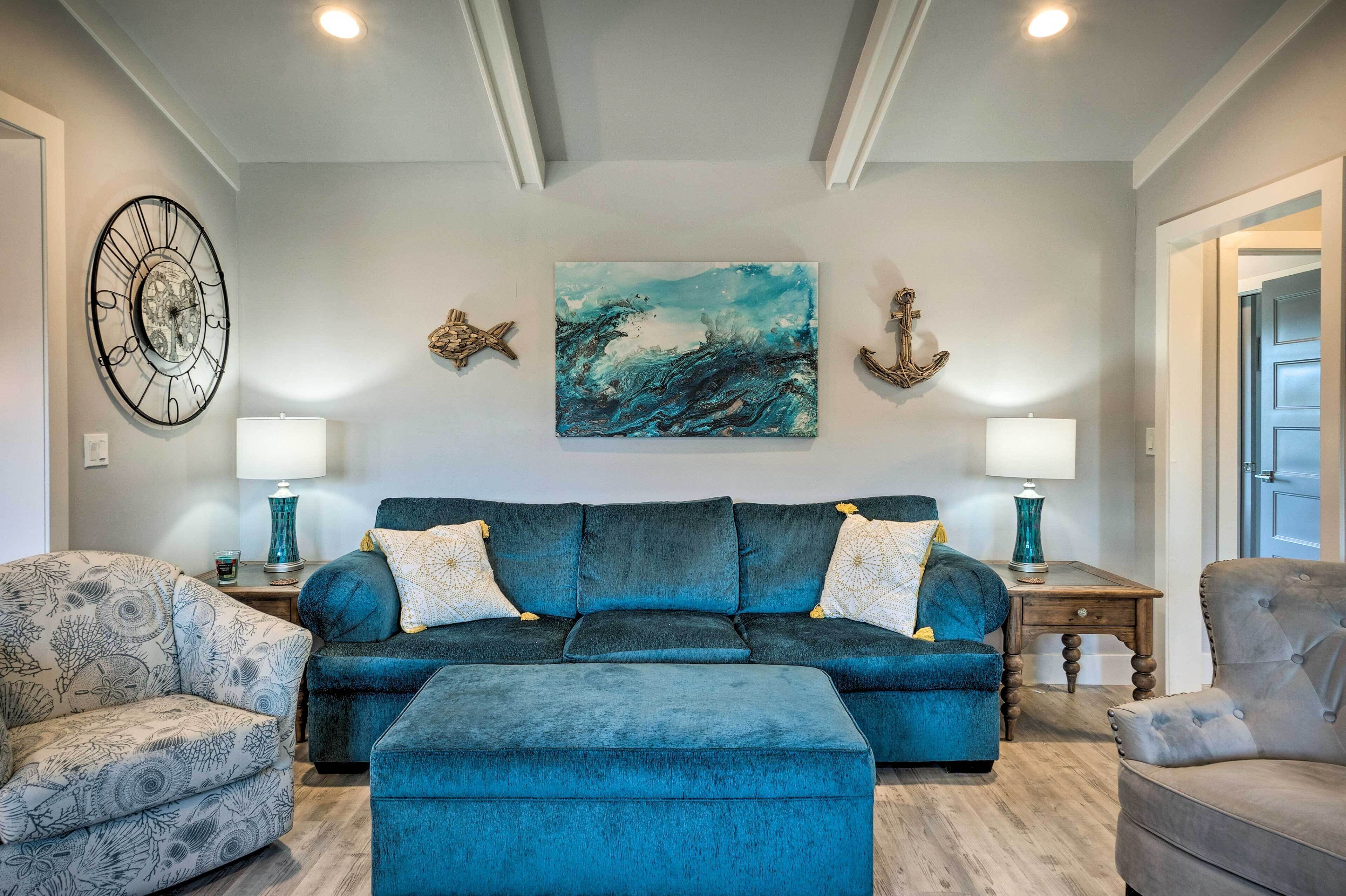 A living room with a blue sofa, patterned armchair, and ottoman. There are two side tables with lamps, a wall clock, and decorative wall art.