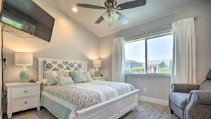 Un dormitorio con cama, mesitas de noche, ventilador de techo, ventana con cortinas y vistas a edificios.