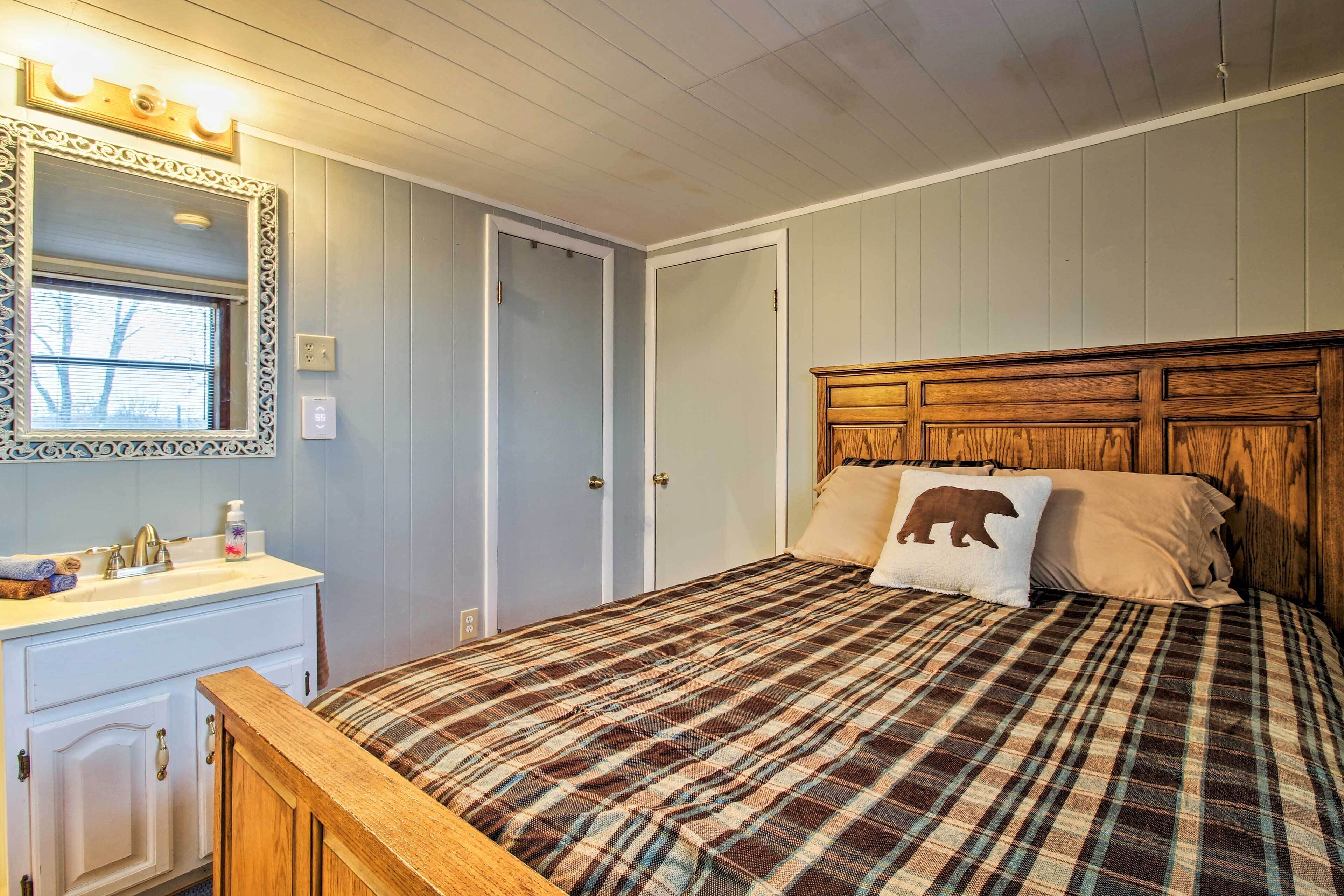 Een slaapkamer met een houten hoofdbord, een geruite spreidekker, een spiegel met verlichting, een wastafel met een wasbak en een raam met jaloezieën.