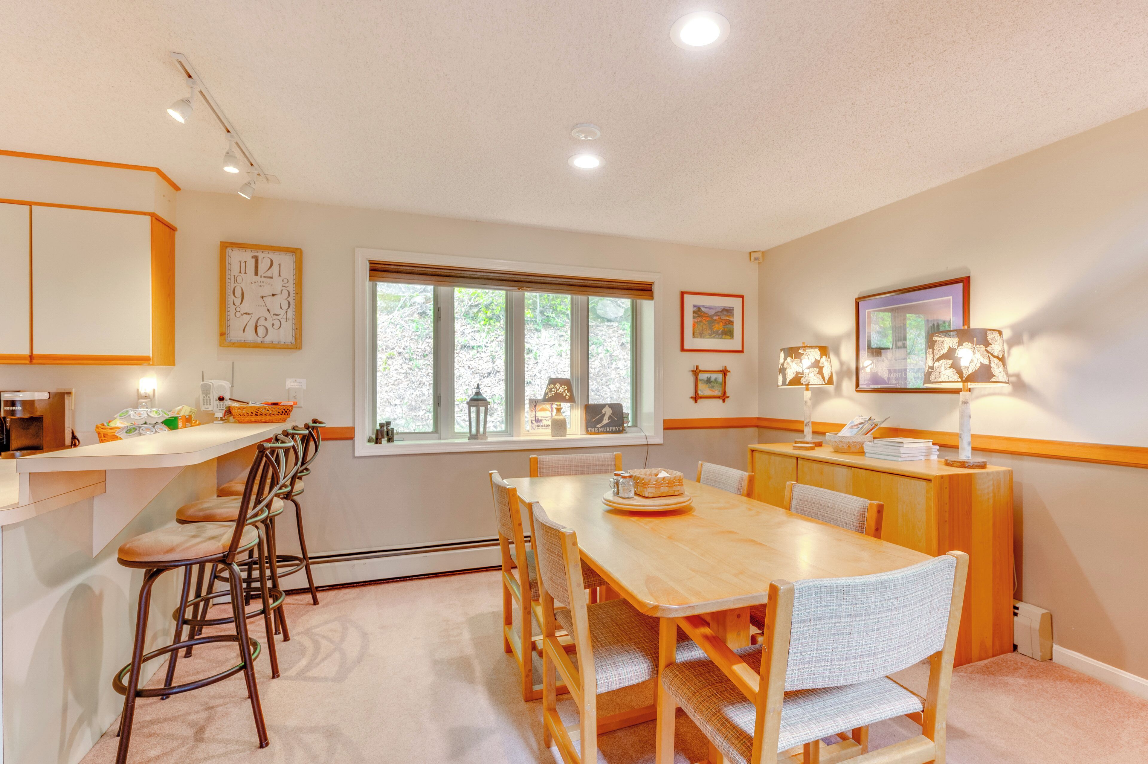 A dining area with a wooden table and chairs, a window with blinds, and a wall-mounted clock.