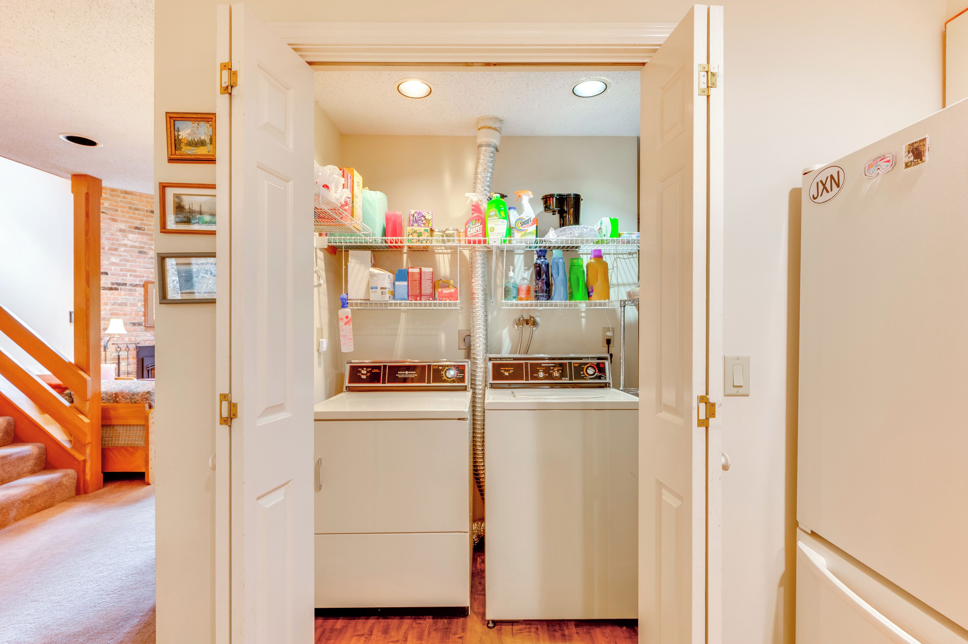 A laundry room with two washers and dryers, a shelf with cleaning supplies, and a wooden staircase leading to another room.
