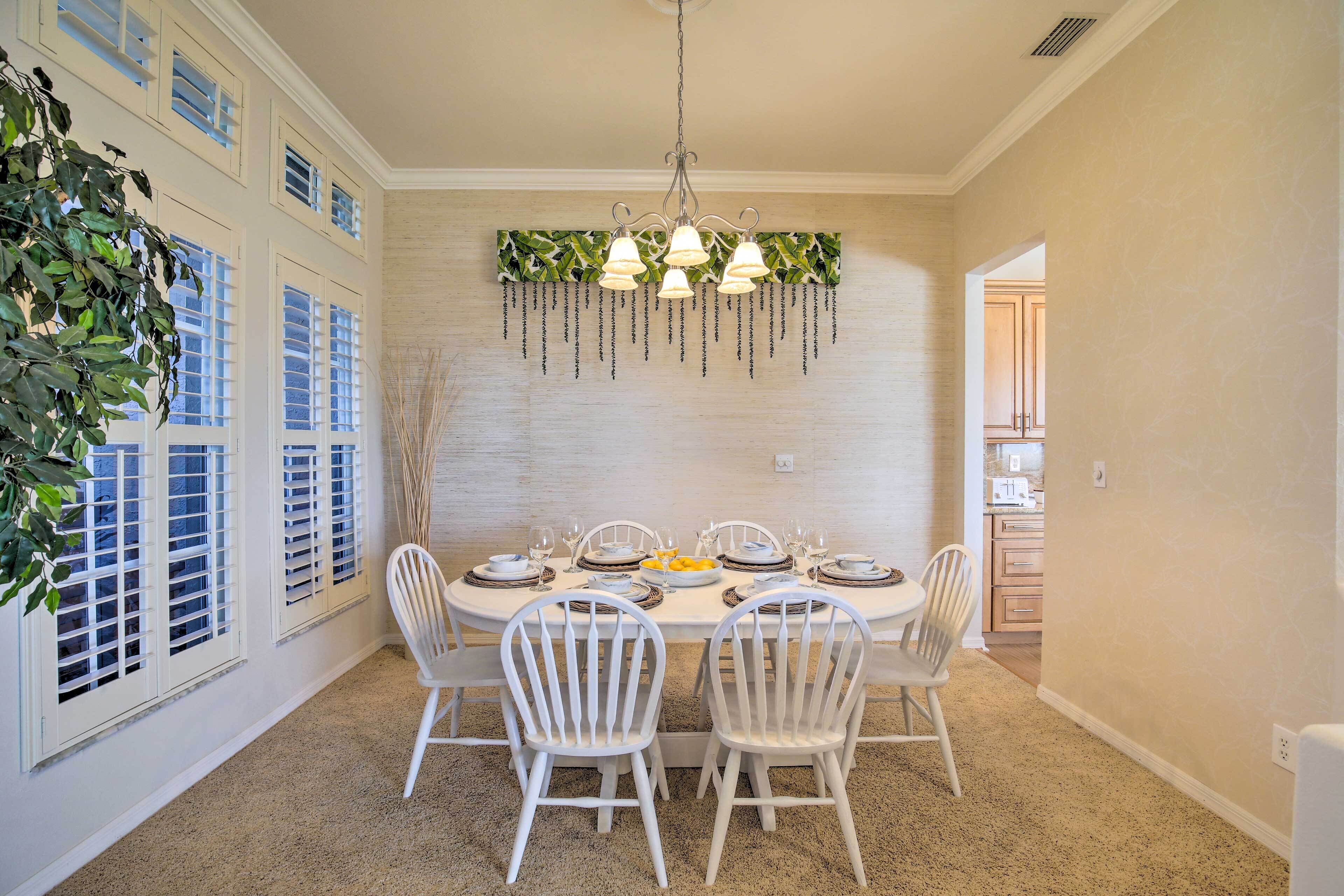 A dining room with a white table set for six, surrounded by white chairs, a hanging light fixture, and a plant on the left.