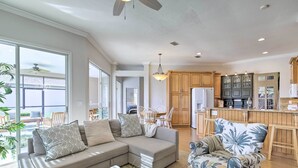 A spacious living room with a sectional sofa, a coffee table, and a patterned armchair. A kitchen with wooden cabinets and a refrigerator is visible in the background. - Bright & Sunny Riverview Oasis w/ Pool & Pond (Riverview)