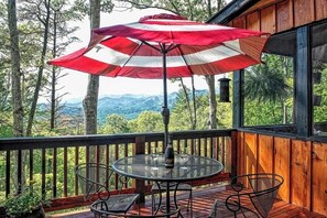 Um deck de madeira com mesa e cadeiras, um guarda-sol e vista para uma floresta e montanhas.
