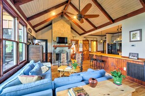 Uma sala de estar ampla com teto alto, vigas de madeira, lareira, televisão e área de cozinha com banquetas.