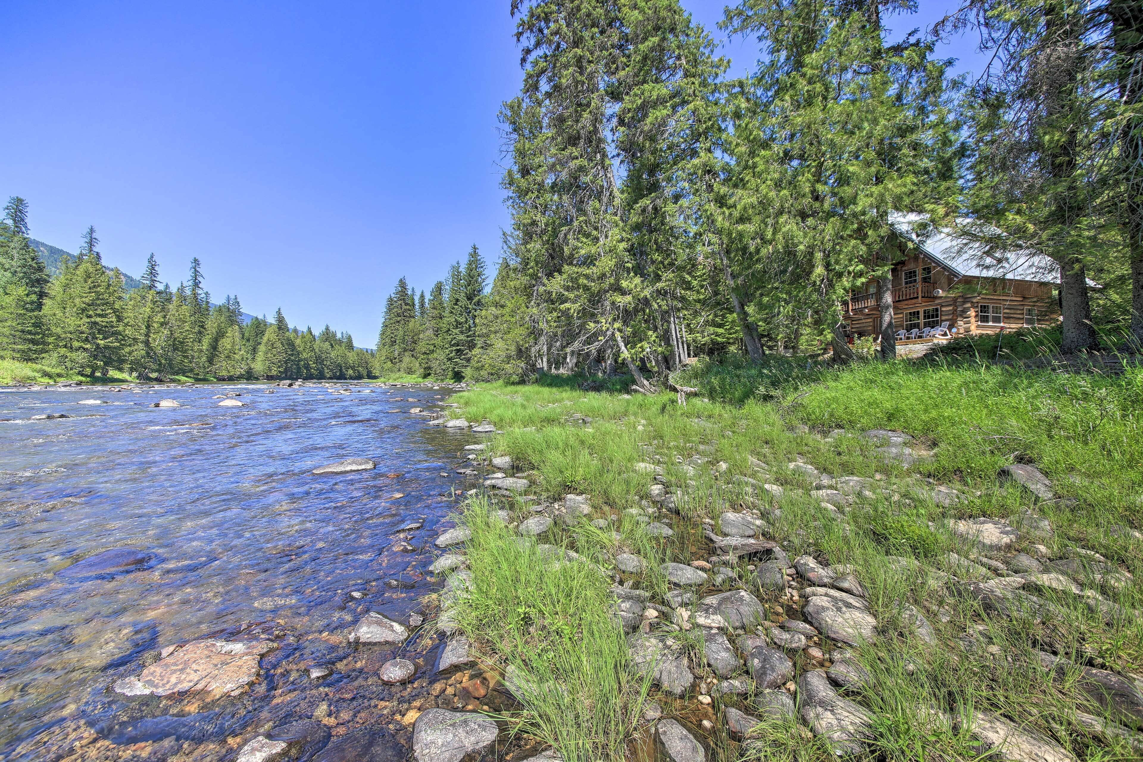 A cabin by a clear river with surrounding trees.