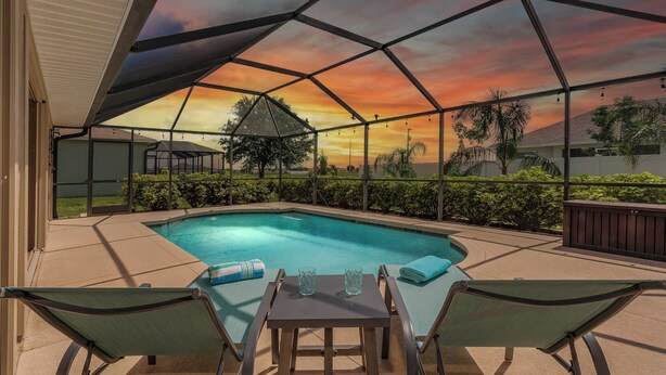 A covered outdoor swimming pool with lounge chairs and a table, surrounded by greenery and a sunset view.