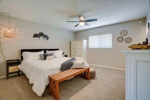 Une chambre à coucher comprenant un lit, un banc en bois, un ventilateur de plafond, une table de chevet, une commode et des œuvres d’art décoratives accrochées au mur.