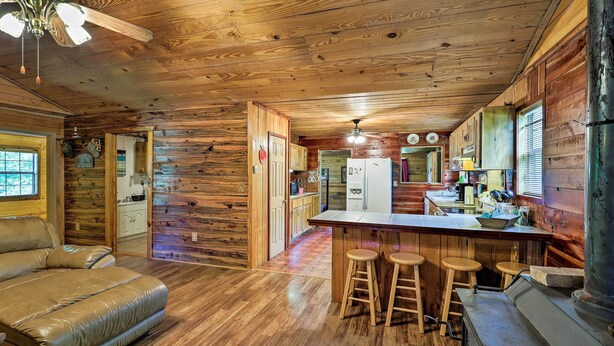 L’intérieur d’une cabane en rondins, avec un plancher en bois, un coin bar doté de tabourets, une cuisine avec des armoires en bois et un ventilateur de plafond.
