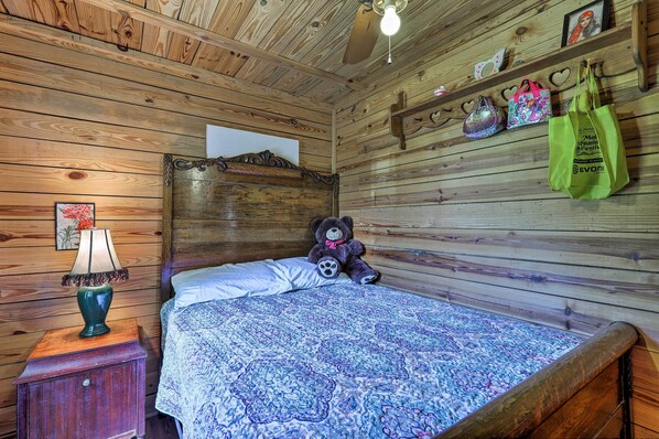 Una habitación con paneles de madera, una cama, un osito de peluche, una lámpara y una estantería con bolsos.