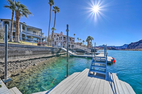 A waterfront property with a wooden dock, clear blue water, and a large building with multiple balconies and palm trees.