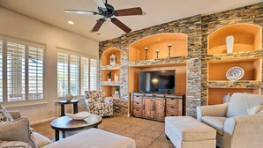 A living room with a stone wall, a ceiling fan, a flat-screen TV, and a fireplace. - 11 Mi to Phoenix Raceway: Luxury Sonoran Oasis! (Goodyear)