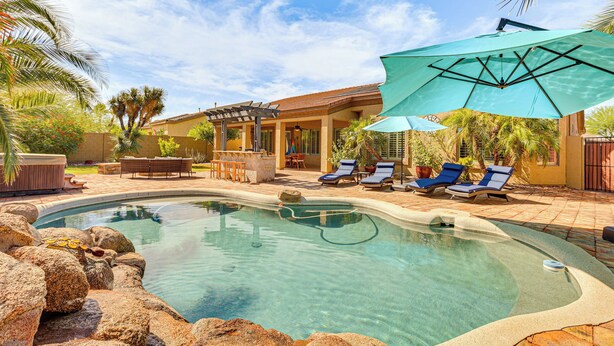 A swimming pool with lounge chairs and umbrellas, surrounded by palm trees and a patio area. - 11 Mi to Phoenix Raceway: Luxury Sonoran Oasis! (Goodyear)