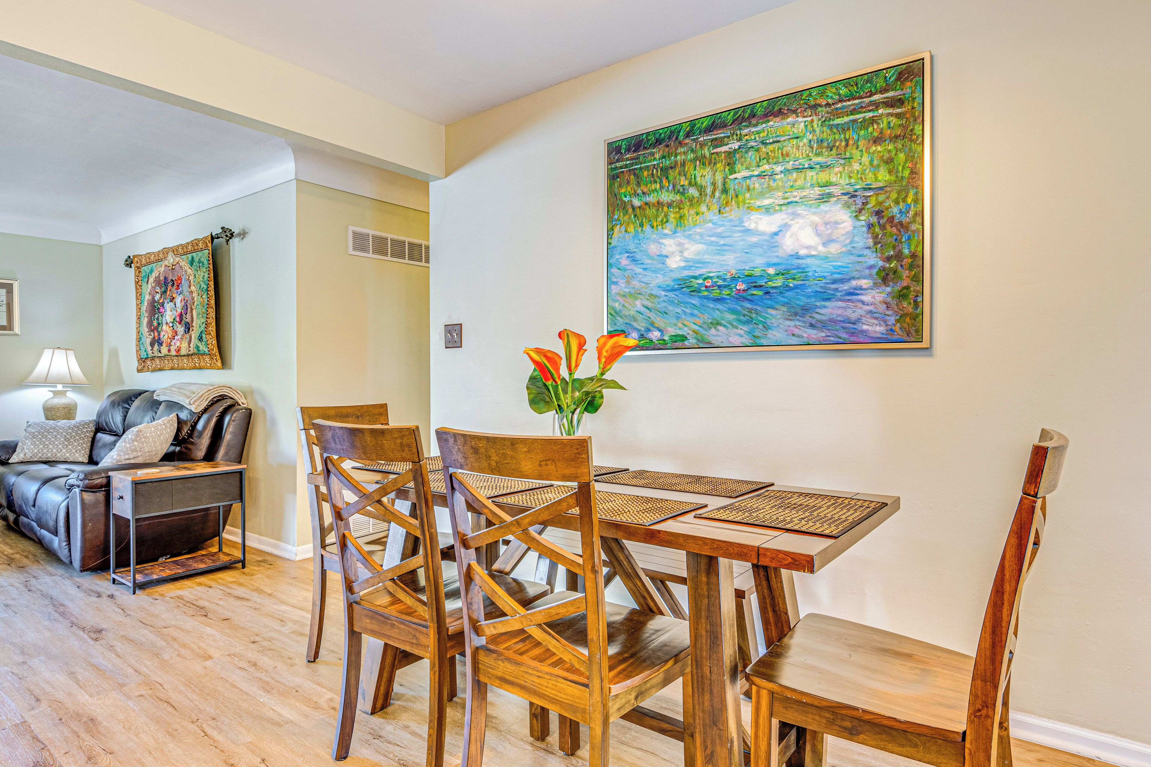 A dining area with a wooden table and chairs, a painting of a landscape, and a sofa in the background.