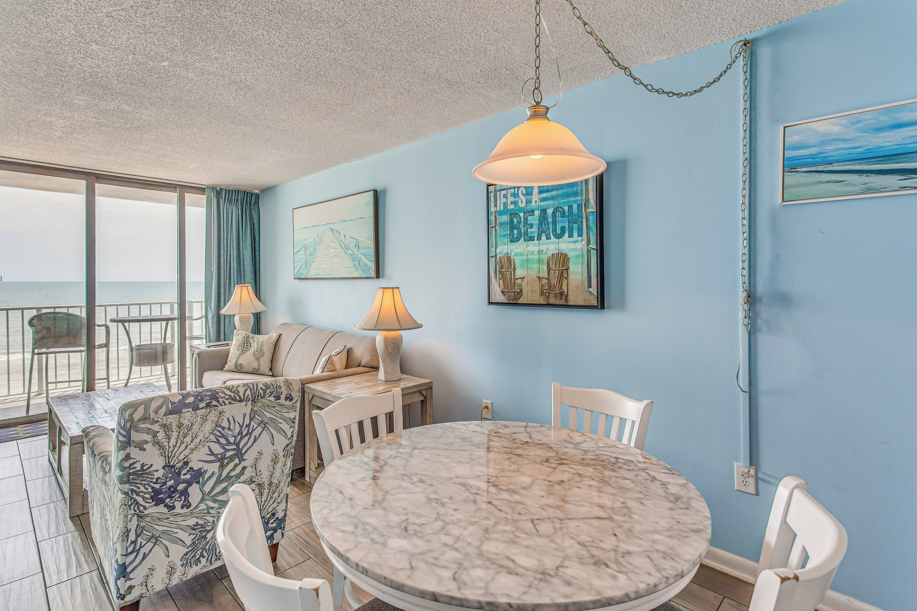 A room with a round marble table, white chairs, a sofa, and a view of the ocean through large windows.