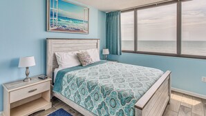 A bedroom with a large bed, a nightstand, a lamp, a painting of a beach, and a view of the sea through a window. - Oceanfront Myrtle Beach Condo With Resort Perks! (Myrtle Beach)