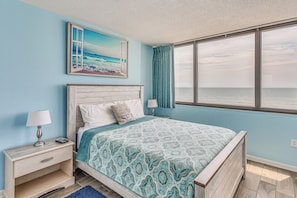 A bedroom with a large bed, a nightstand, a lamp, a painting of a beach, and a view of the sea through a window.