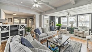 A spacious living room with a grey sofa, striped armchairs, and a coffee table. There are ceiling fans, a kitchen area in the background, and large windows providing natural light.