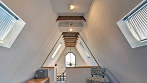 Une petite pièce mansardée aux poutres en bois inclinées, avec un petit bureau, une chaise à motifs et une fenêtre équipée de stores.