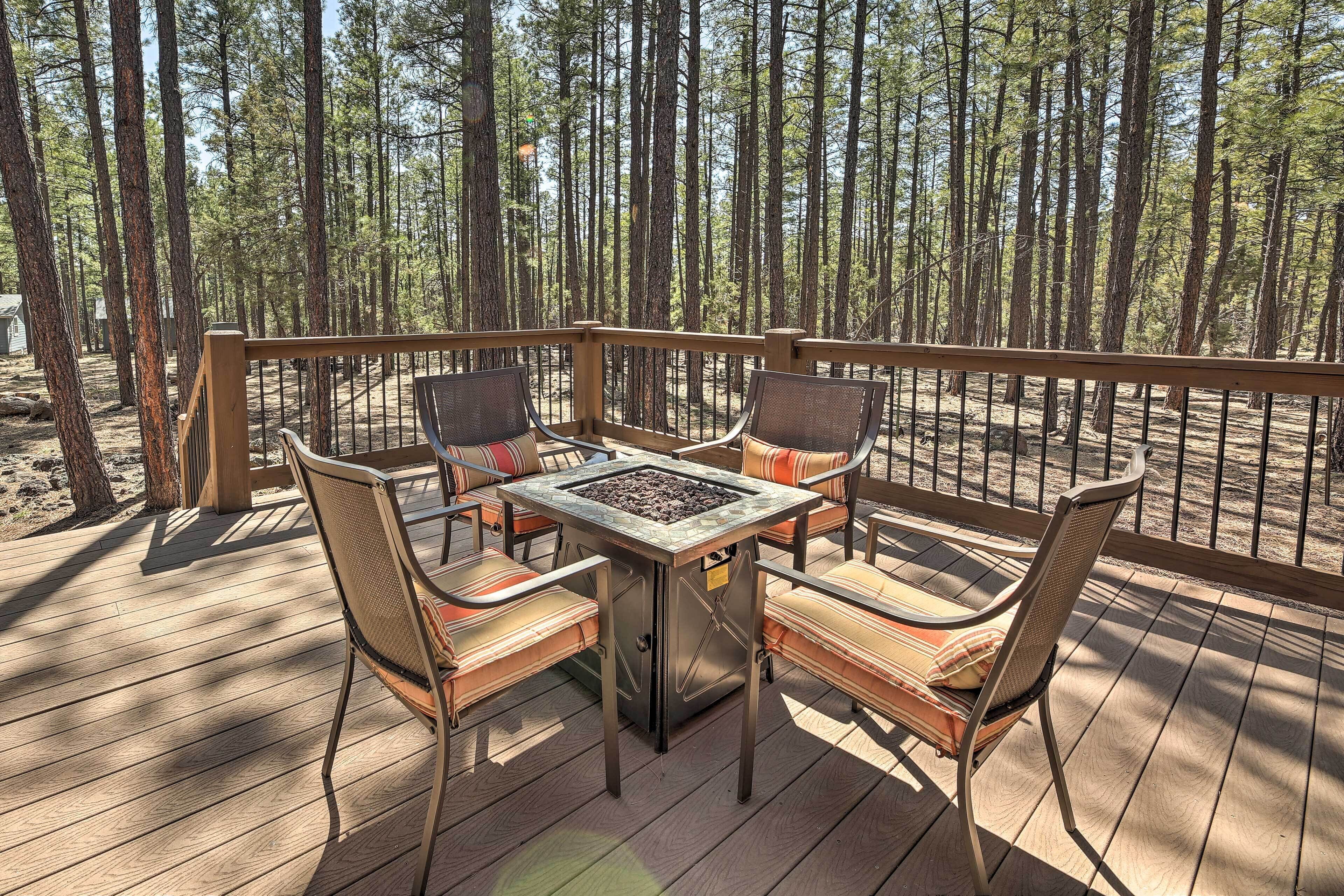 Eine Terrasse mit Feuertisch und vier Stühlen, umgeben von hohen Bäumen.