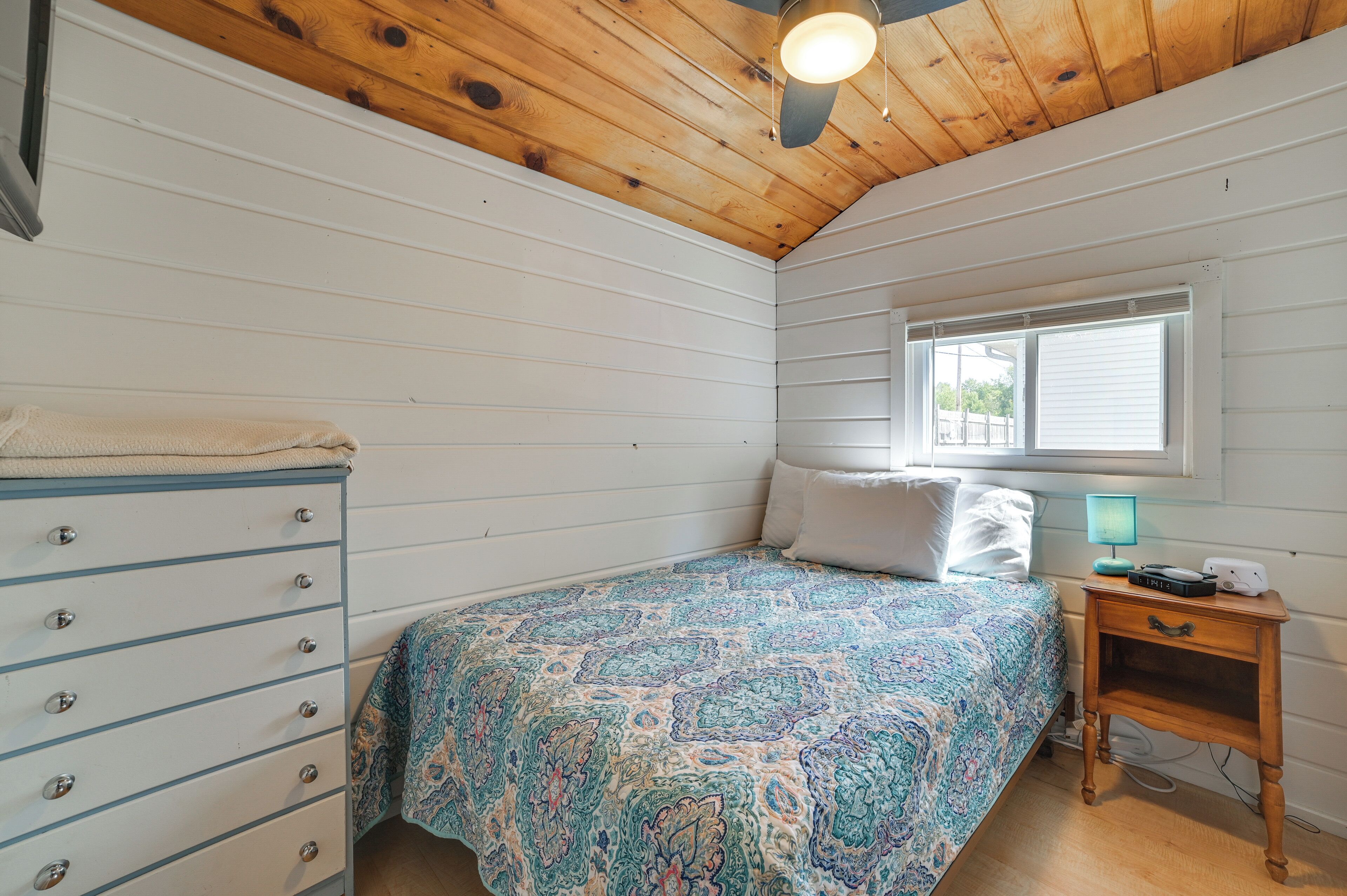 Un dormitorio con una cama, un aparador, una mesita de noche, una ventana y un ventilador de techo.