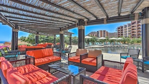 Un espace extérieur aménagé, abrité et doté de mobilier en osier et de coussins orange, offrant une vue sur une piscine et les bâtiments de l’hôtel.