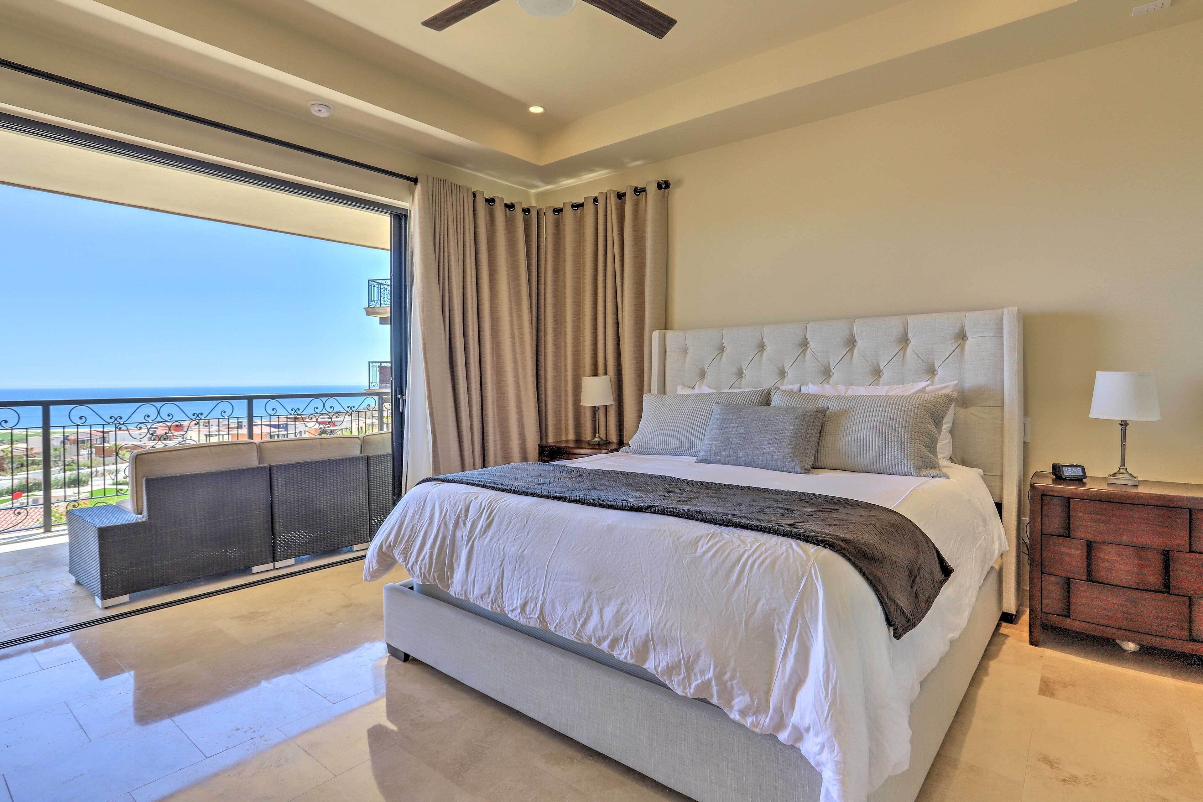 A bedroom with a large bed, a nightstand, a dresser, and a balcony with a view of the sea.