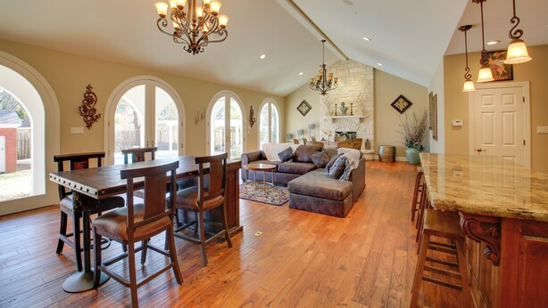 A spacious living area with a wooden dining table, bar stools, a sofa, and a kitchen area with a stone fireplace. - Hot Springs Home With Pool - 1/2 Mile to Oaklawn! (Hot Springs)