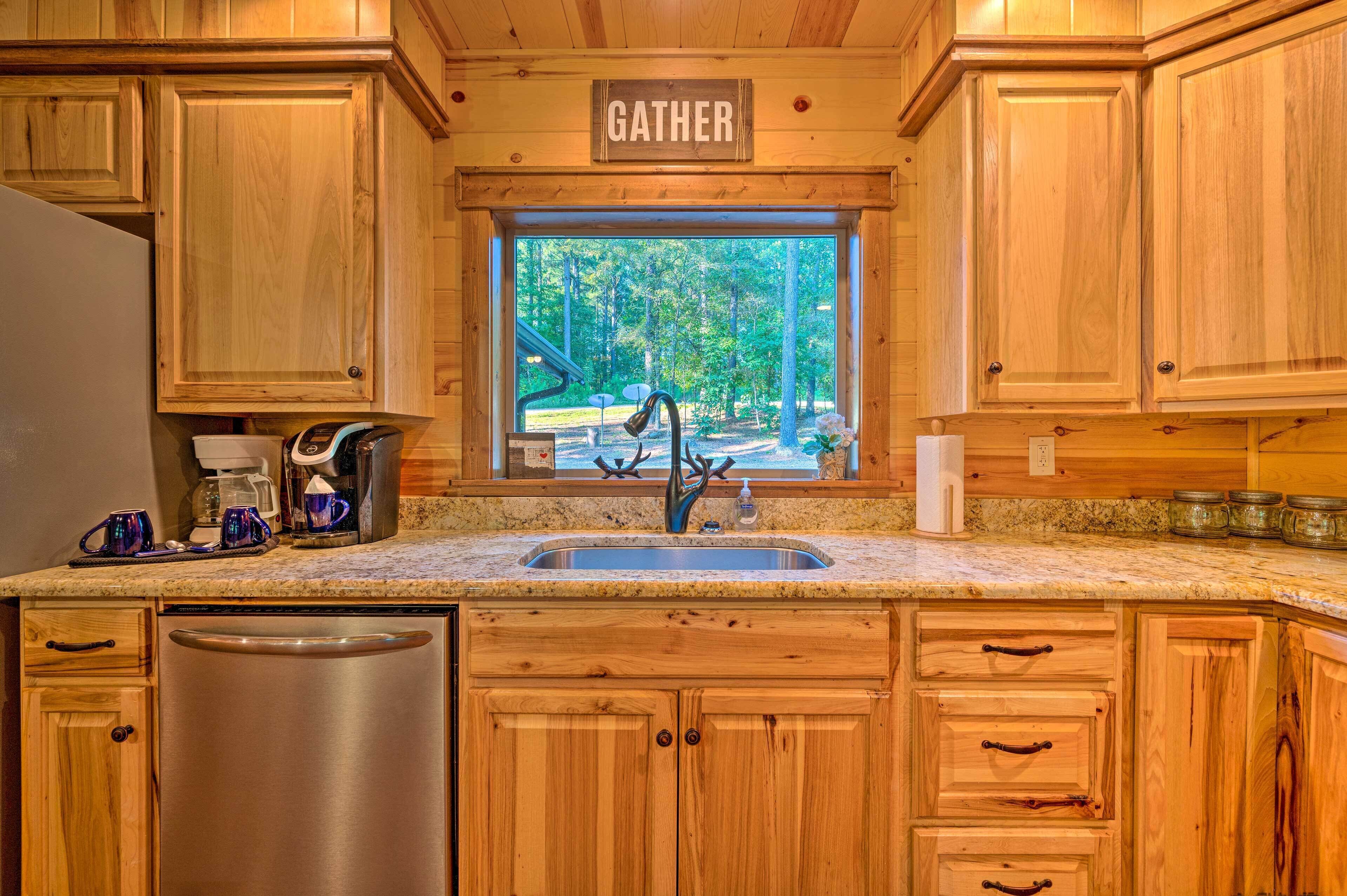 A wooden kitchen with a granite countertop, stainless steel dishwasher, and a window with a view of trees.