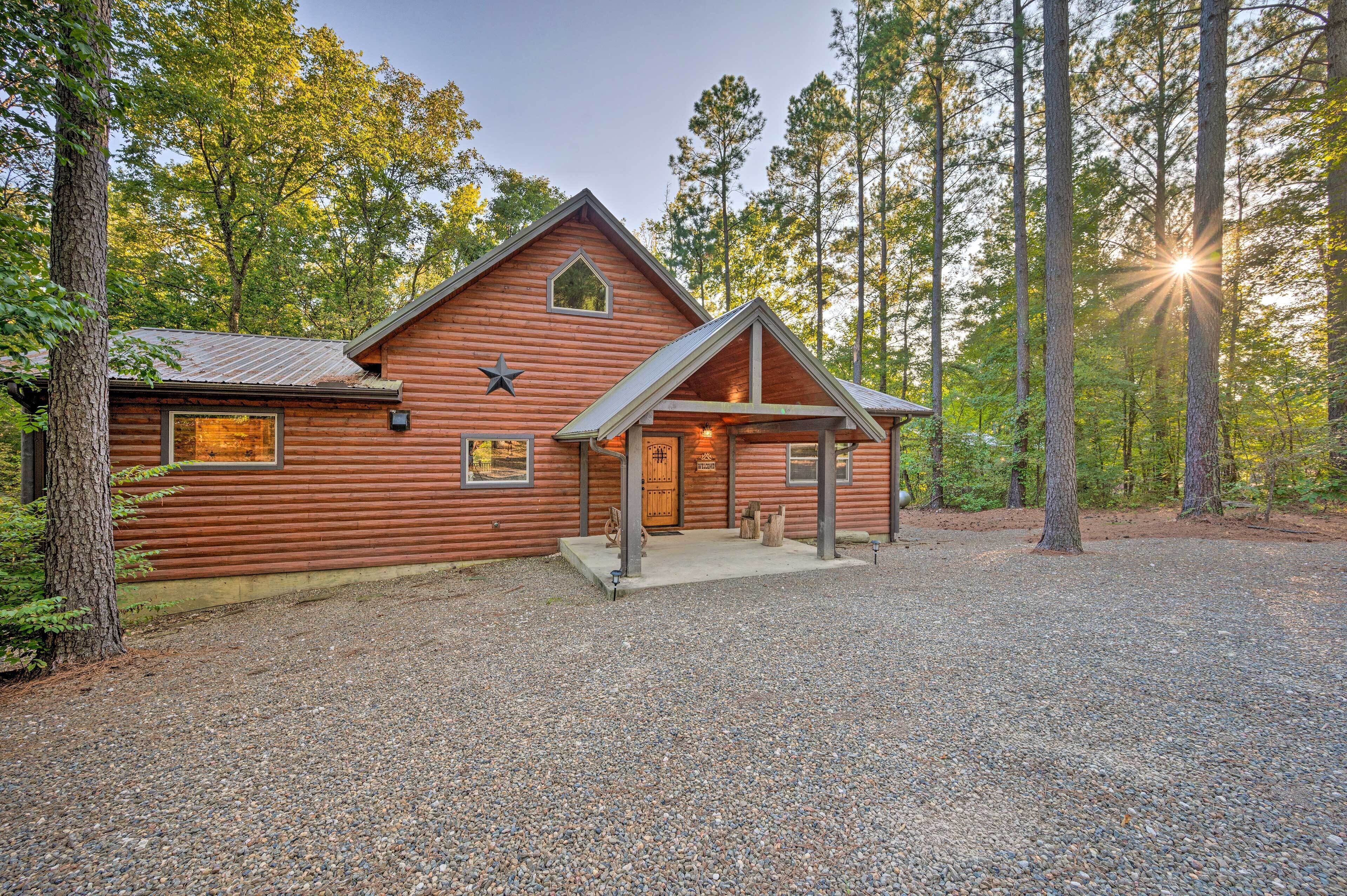 A log cabin with a porch, surrounded by trees and a gravel driveway.