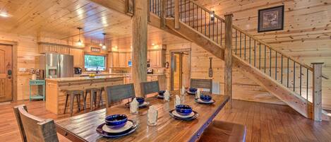 A spacious wooden cabin with a large dining table set for six, a kitchen area, and a staircase.