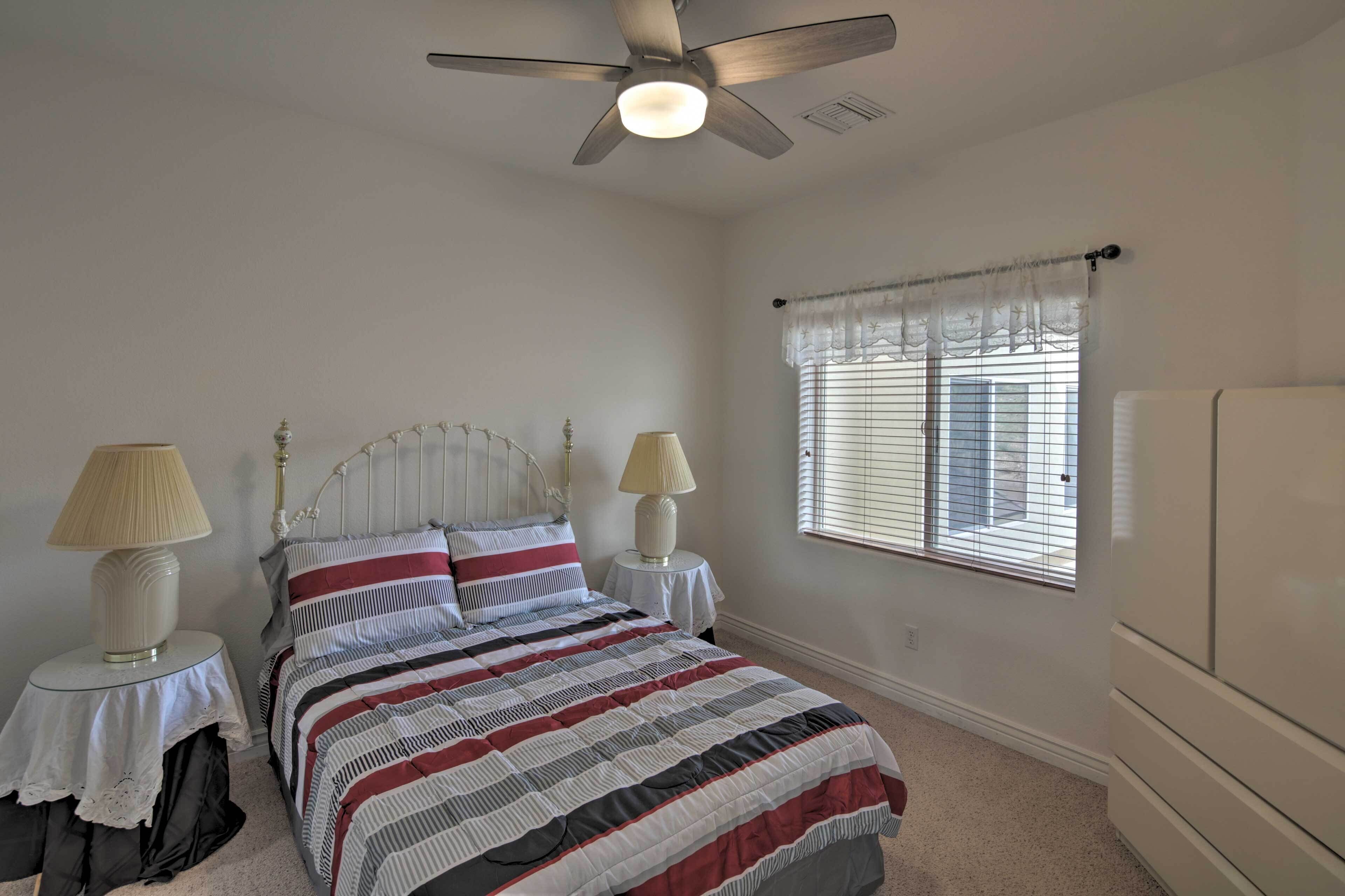 A bedroom with a bed, two bedside lamps, a ceiling fan, a window with blinds, and a white wardrobe.