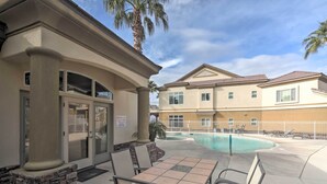 Un espace aménagé au bord de la piscine, avec une table et des chaises, un palmier, et un bâtiment à la façade beige, avec une piscine en arrière-plan.