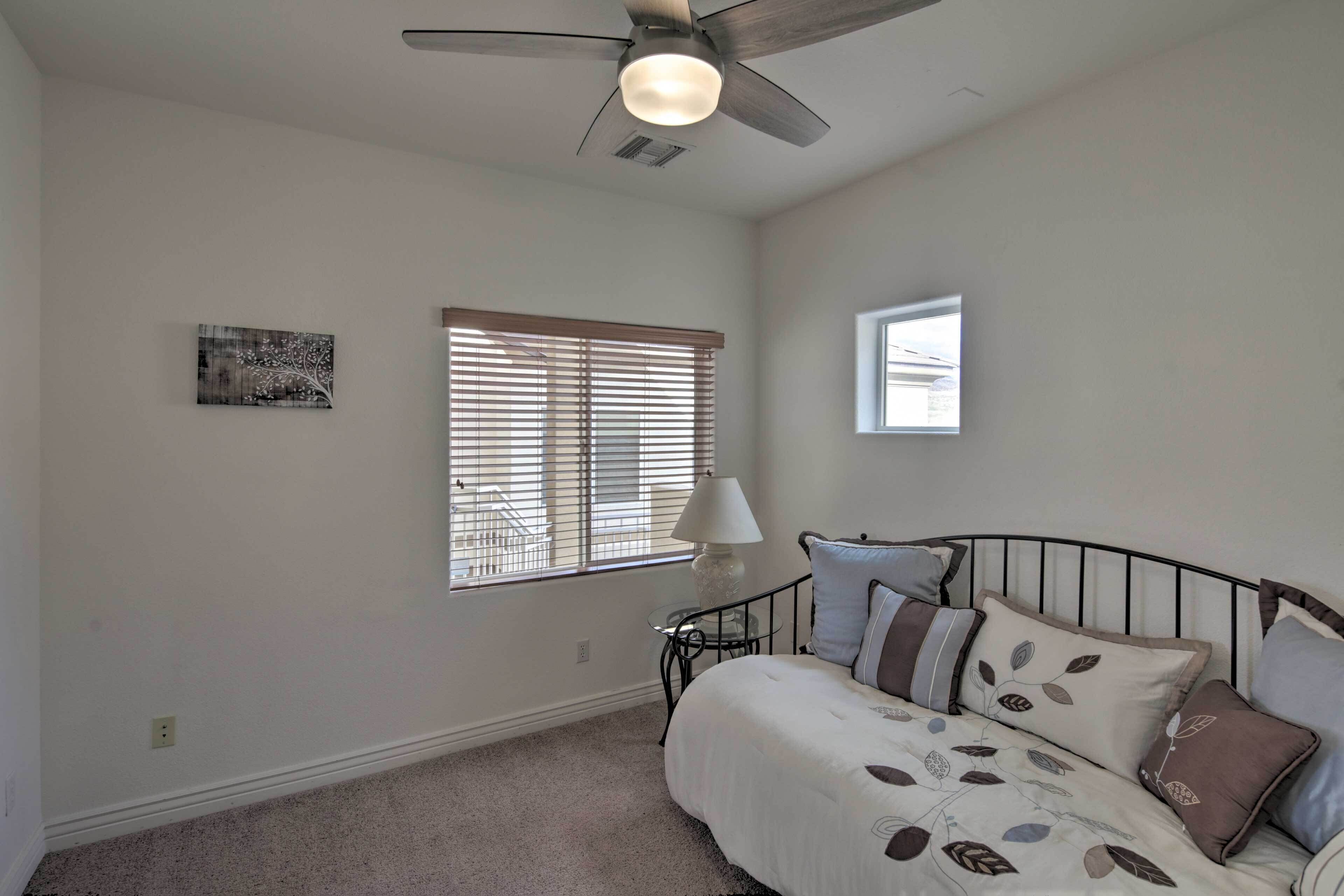 A bedroom with a bed, a ceiling fan, a window with blinds, and a small table with a lamp.