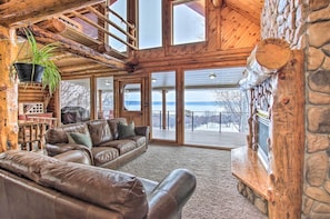 A cozy cabin interior with a leather sofa, a stone fireplace, and large windows offering a view of the outdoors.