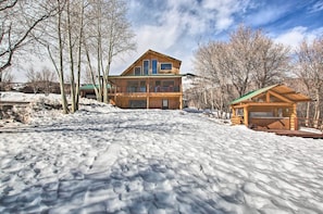 A wooden cabin with a large deck and a separate garage, surrounded by snow-covered ground and leafless trees.