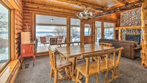 A log cabin interior with a wooden dining table, benches, a fireplace, and large windows offering a view of the outdoors.