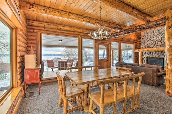 A log cabin interior with a wooden dining table, benches, a fireplace, and large windows offering a view of the outdoors.