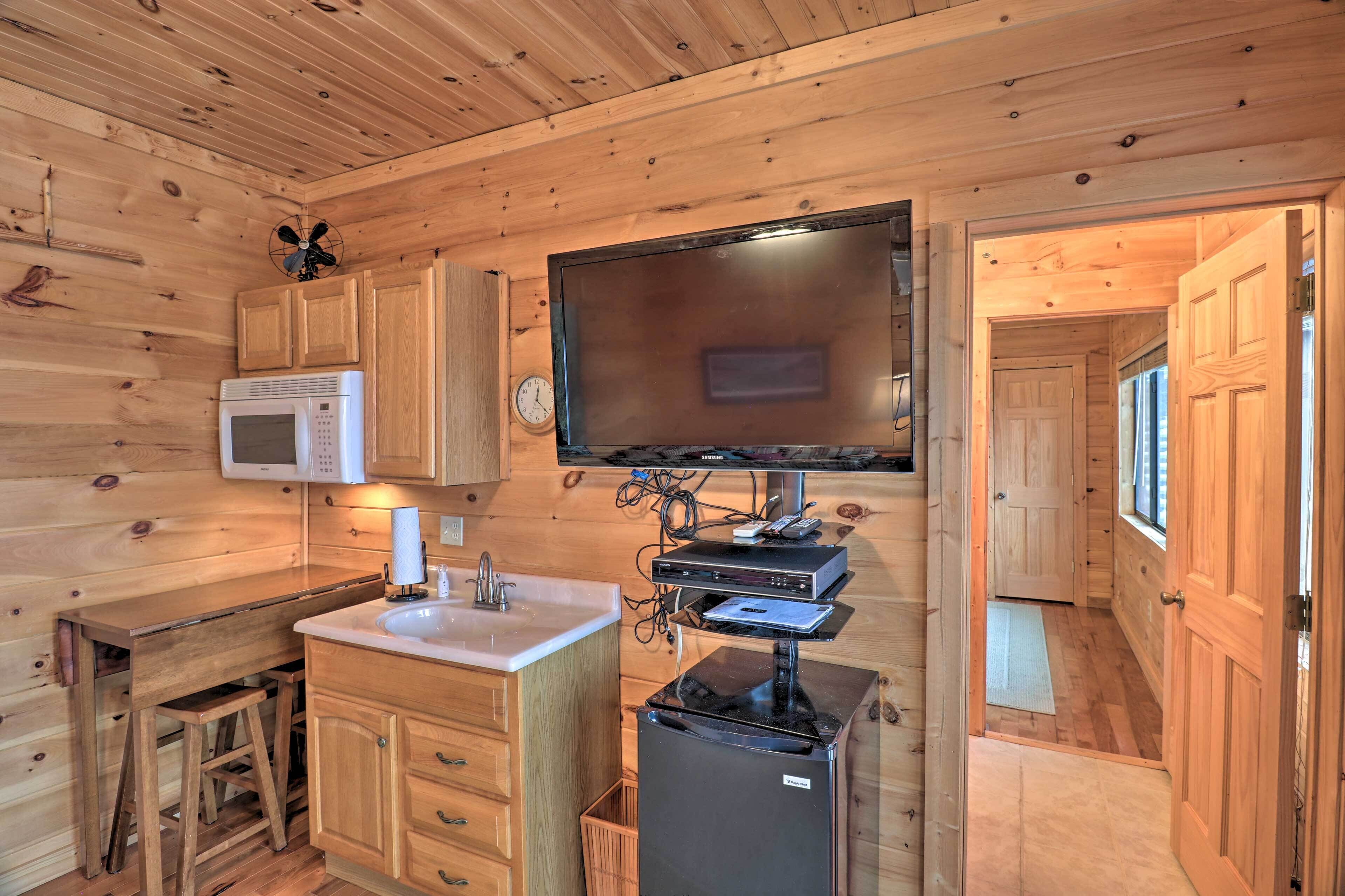 A compact kitchen with wooden walls, a microwave, a sink, a refrigerator, and a flat-screen TV mounted on the wall.