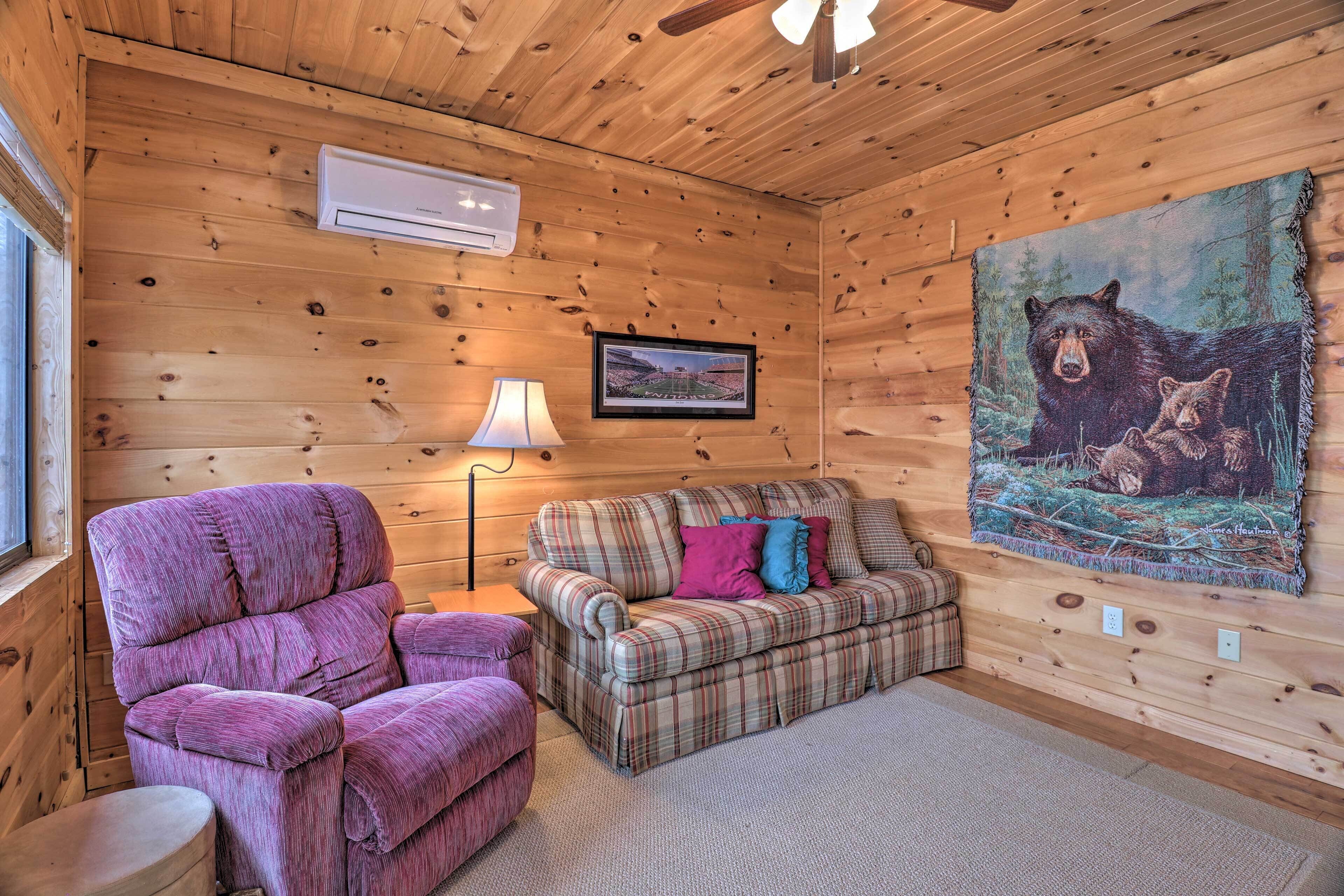 A cozy cabin interior with a plaid sofa, a purple armchair, and a bear-themed painting on the wall.