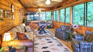 Ein gemütliches Blockhaus-Interieur mit Holzvertäfelung, einer Couch, einem Tisch und einem großen Fenster mit Blick ins Grüne.