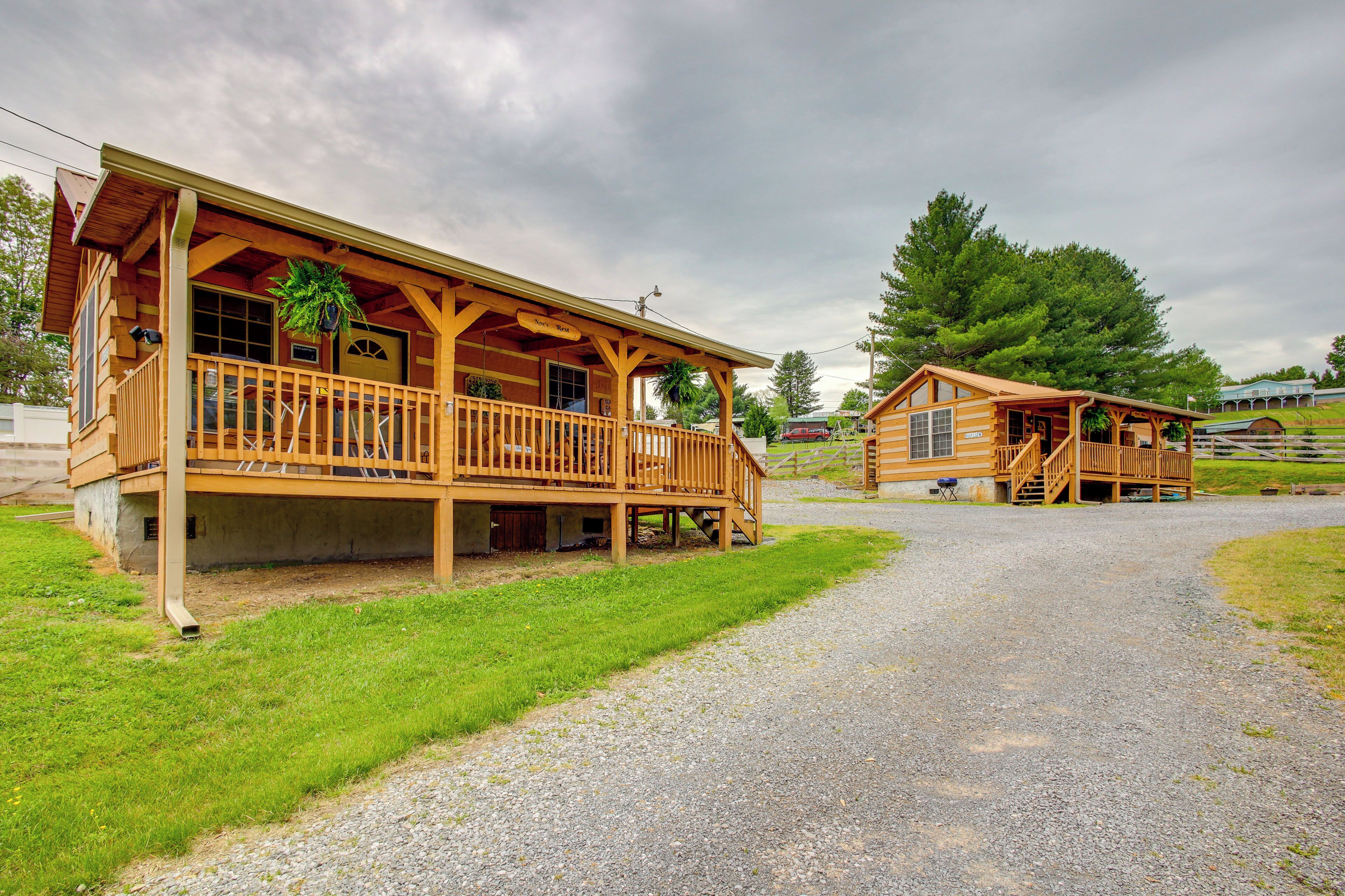 A wooden lodge with a porch and a covered deck area, surrounded by a gravel driveway and grassy areas.