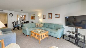 A living room with a sectional sofa, a glass coffee table, a TV on a stand, and a dining area with chairs and a table. - Central Cocoa Beach Condo w/ View, Steps to Beach! (Cocoa Beach)