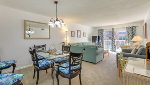 A living room with a dining table, chairs, a sofa, and a balcony with curtains. - Central Cocoa Beach Condo w/ View, Steps to Beach! (Cocoa Beach)