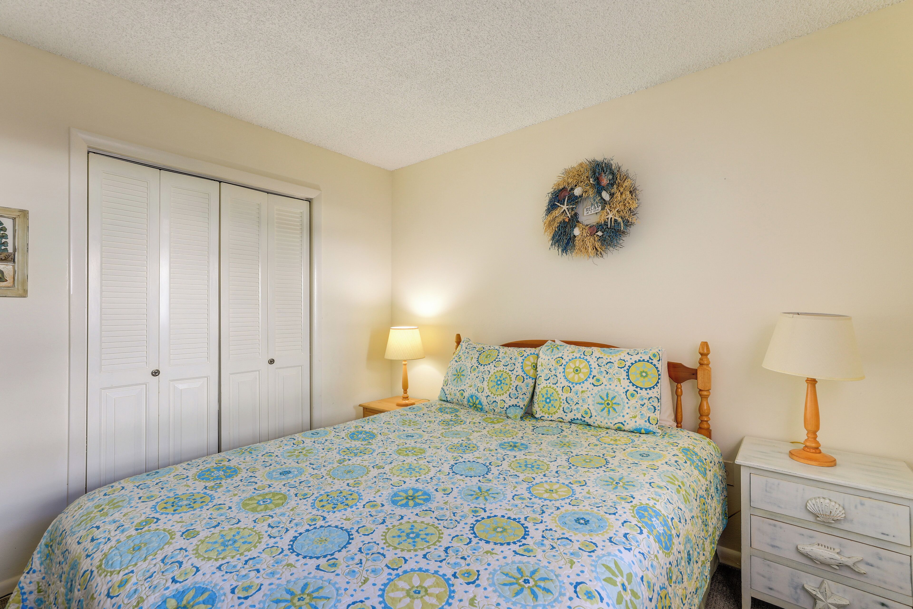 A bedroom with a bed, two nightstands, a wall-mounted lamp, a door, and a decorative wreath.
