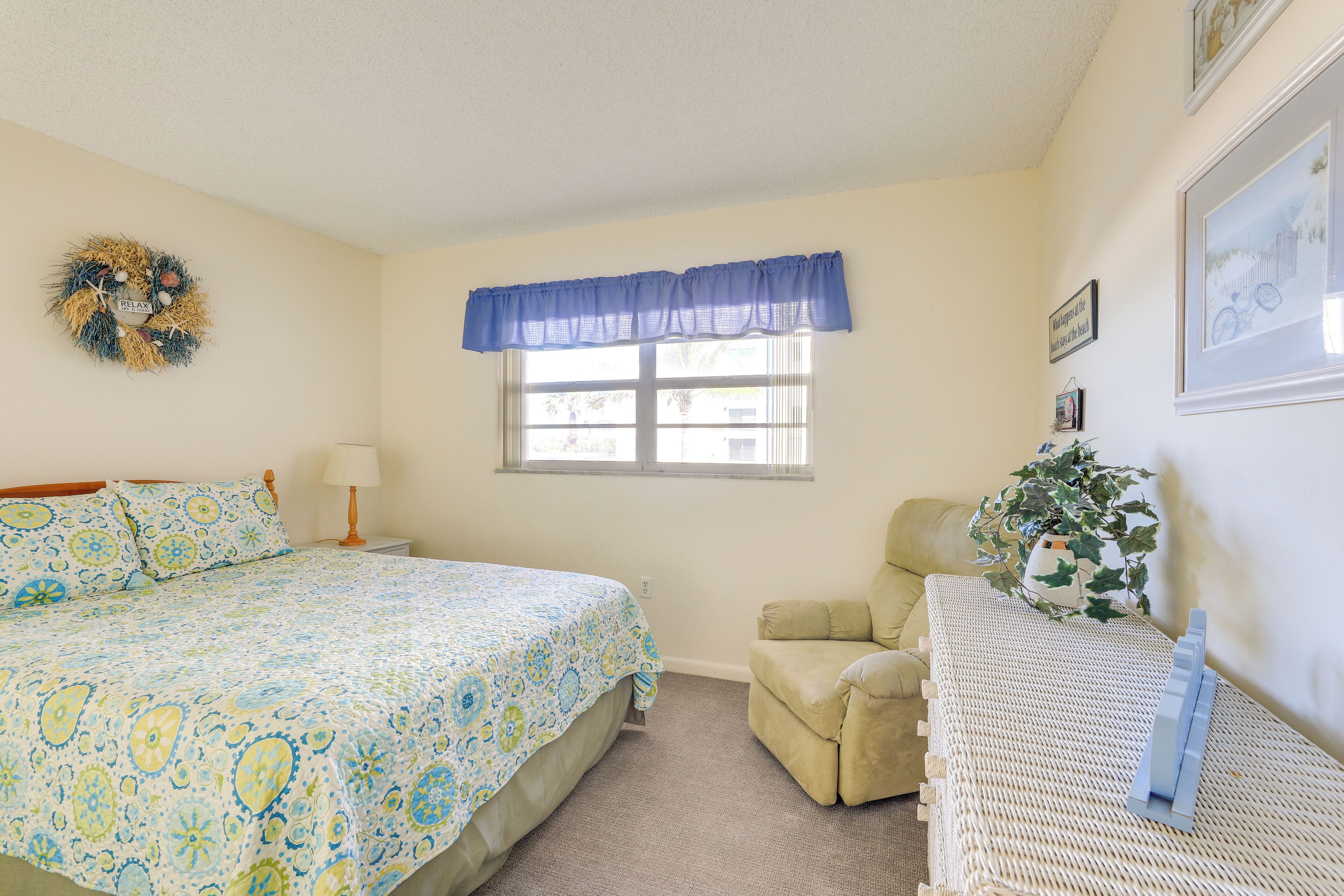 A bedroom with a bed, a chair, a window with a blue valance, a wall-mounted wreath, and a framed picture.