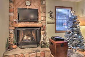 A cozy living room with a stone fireplace, a television on a wooden shelf, a Christmas tree, and a 'WELCOME' sign. - Cozy Angel Fire Condo < Half Mi to Resort! (Angel Fire)