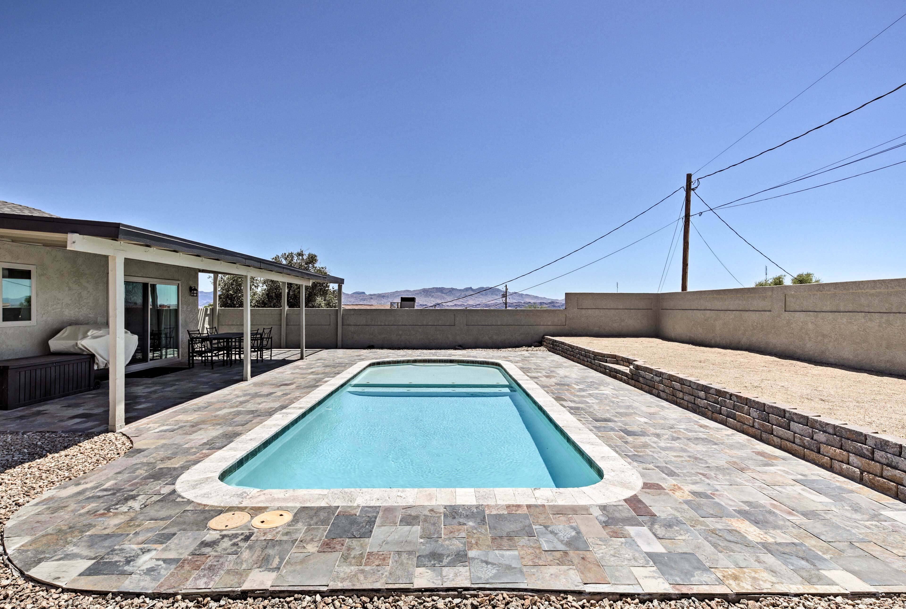 A rectangular swimming pool with a stone tile deck, a covered patio area with outdoor furniture, and a view of mountains in the background.