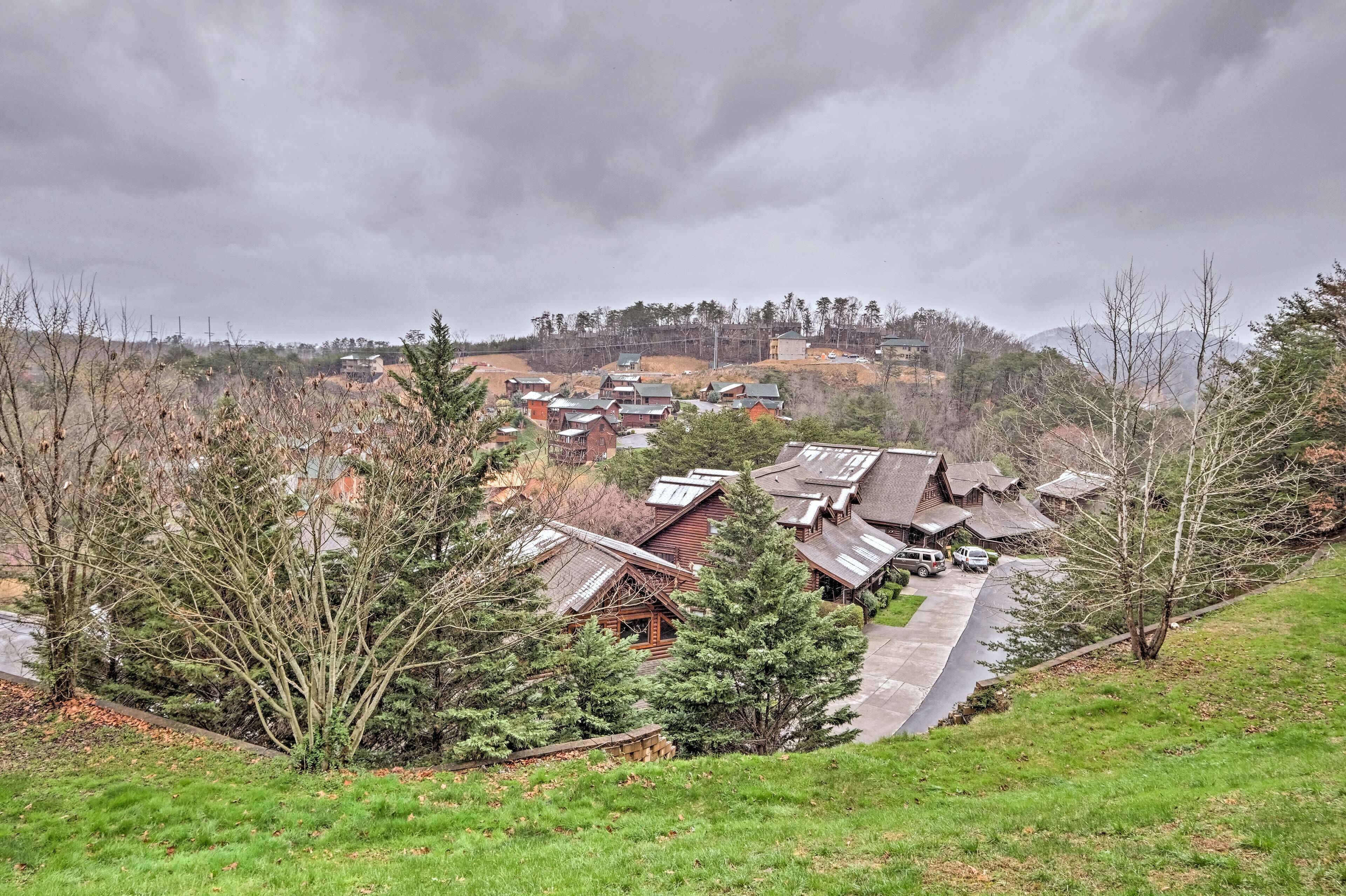 A residential area with houses on a hillside, surrounded by trees and a cloudy sky.