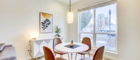 Un comedor moderno con una mesa redonda, cuatro sillas y una lámpara colgante. Un armario blanco con diseño geométrico está contra la pared, y hay una obra de arte enmarcada encima.